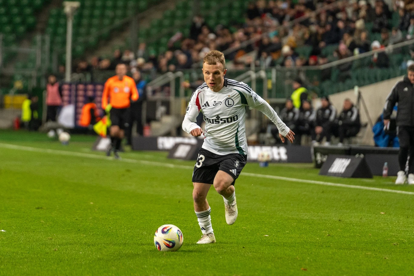 Piłkarz w białym stroju z logotypem Legii Warszawa prowadzi piłkę na zielonej murawie podczas meczu, w tle widoczni sędzia, ławka rezerwowych oraz kibice na trybunach.