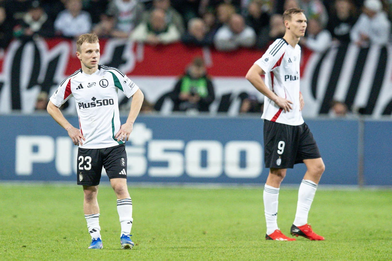 Dwóch piłkarzy Legii Warszawa w białych koszulkach stoi na murawie stadionu podczas meczu, obaj sprawiają wrażenie skupionych lub zawiedzionych, w tle kibice oraz baner reklamowy.