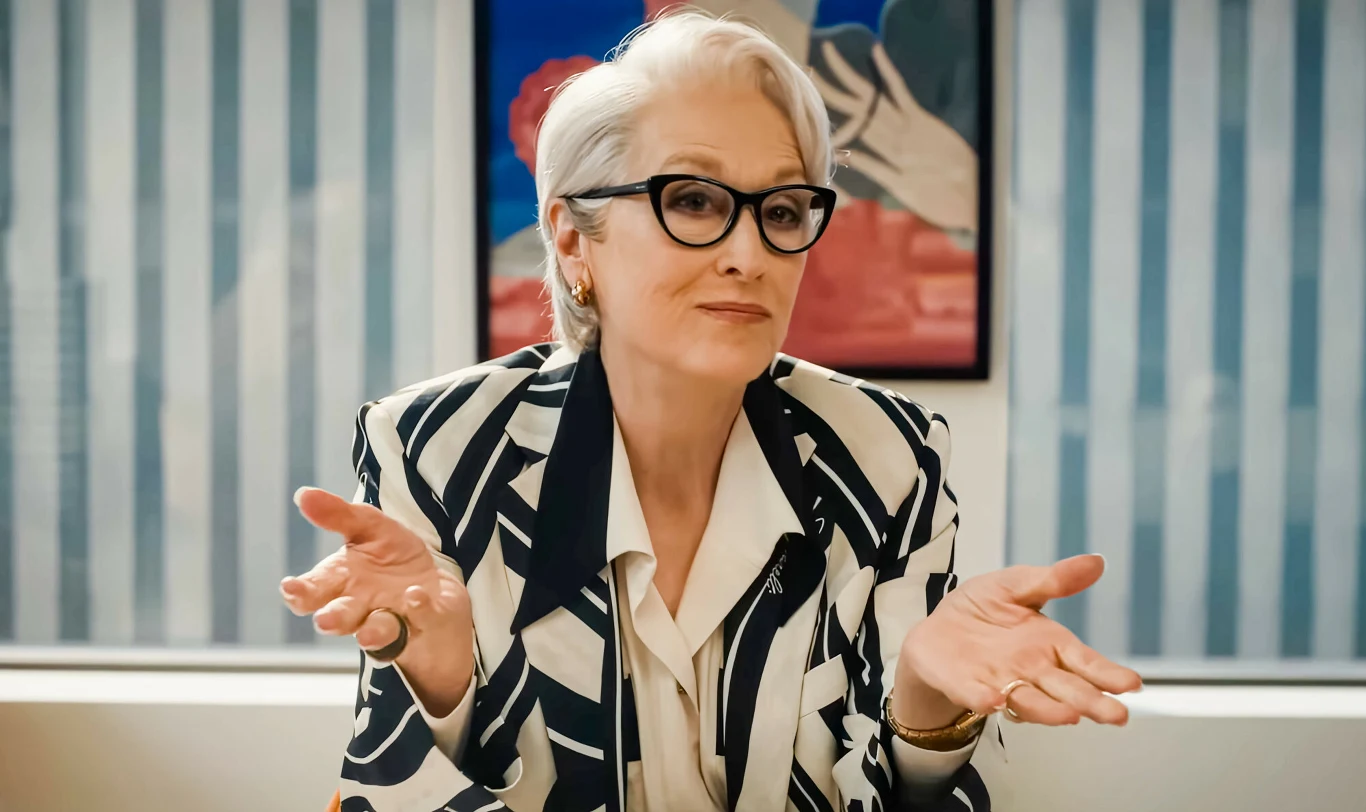 Meryl Streep w filmie "Diabeł ubiera się u Prady 2" kobieta w okularach, o krótkich siwych włosach, ubrana w elegancki żakiet w czarno-białe wzory, siedzi przy biurku z rozłożonymi rękami i neutralnym wyrazem twarzy; w tle widoczny obraz z motywem abstrakcyjnym oraz pionowe jasnoniebieskie żaluzje