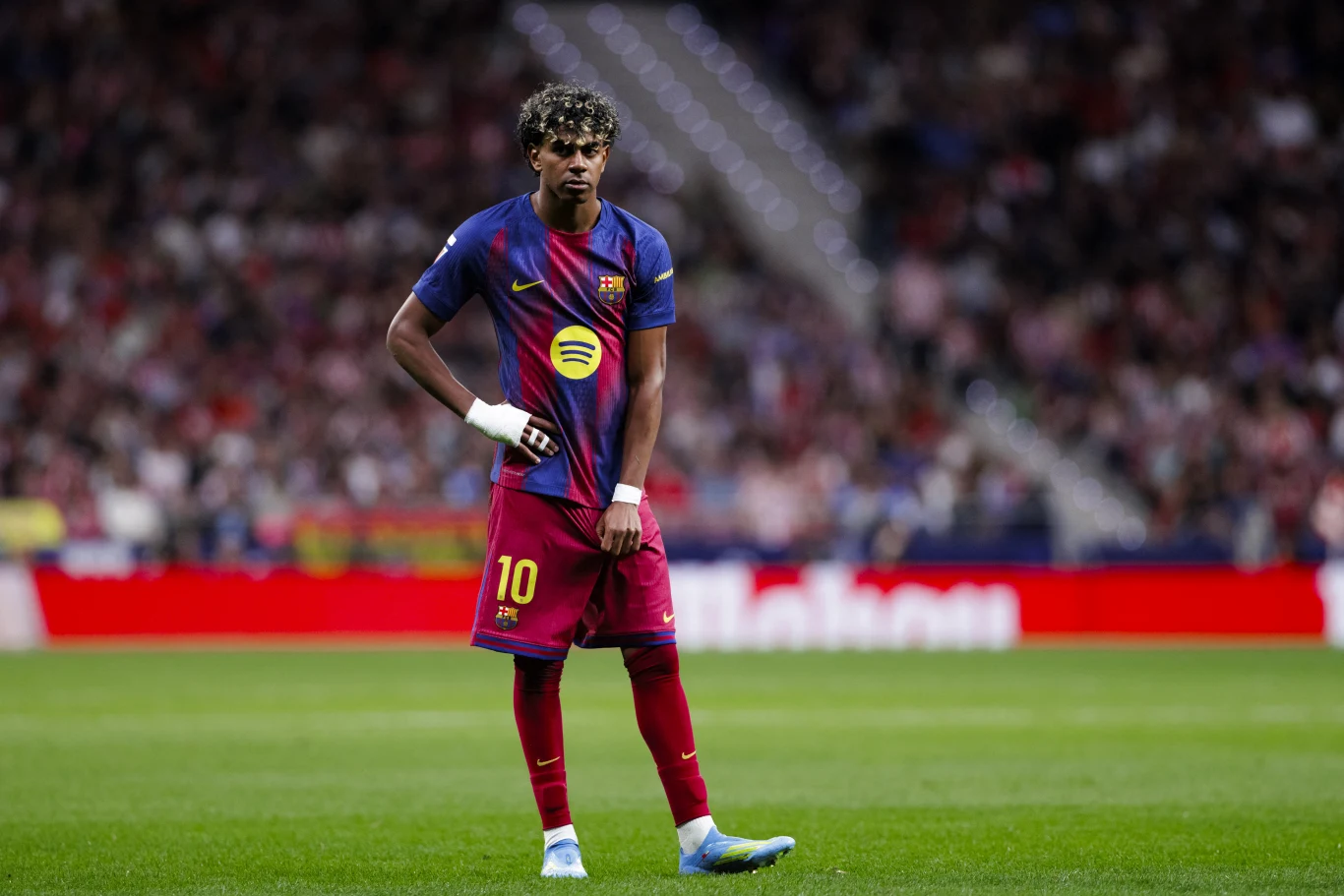 piłkarz w bordowo-granatowej koszulce z numerem 10 i opaską na nadgarstku stoi na murawie stadionu, za nim rozmazana publiczność na trybunach