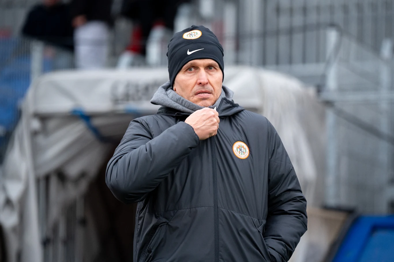 mężczyzna w ciemnej kurtce i czapce z logo klubu sportowego, zaciągający zamek przy szyi w chłodnej scenerii stadionu