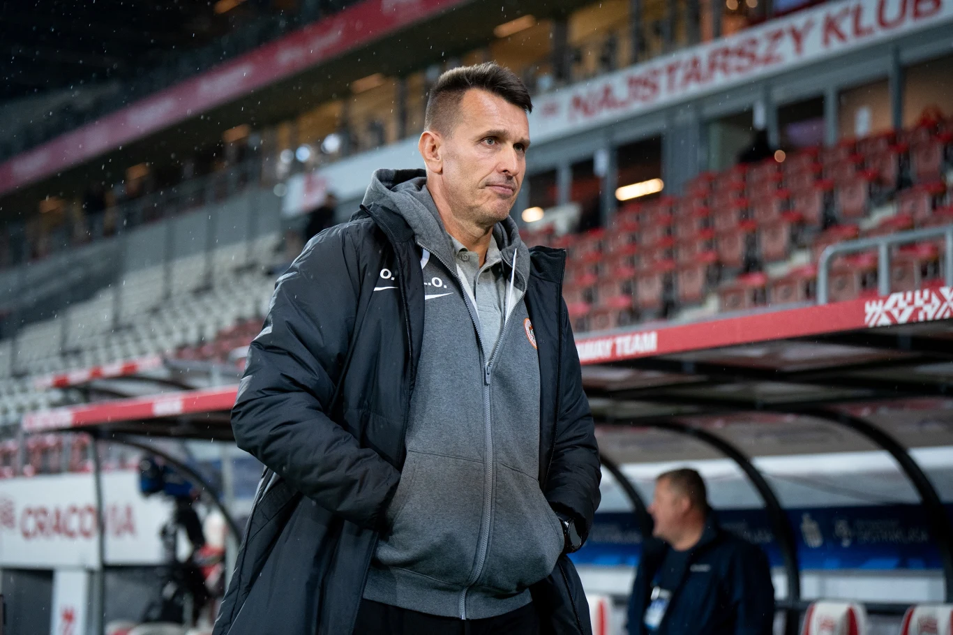 mężczyzna w sportowej kurtce i szarej bluzie stoi przed ławką rezerwowych na stadionie piłkarskim, spoglądając w stronę boiska, w tle widoczne puste trybuny oraz kilka osób na ławce