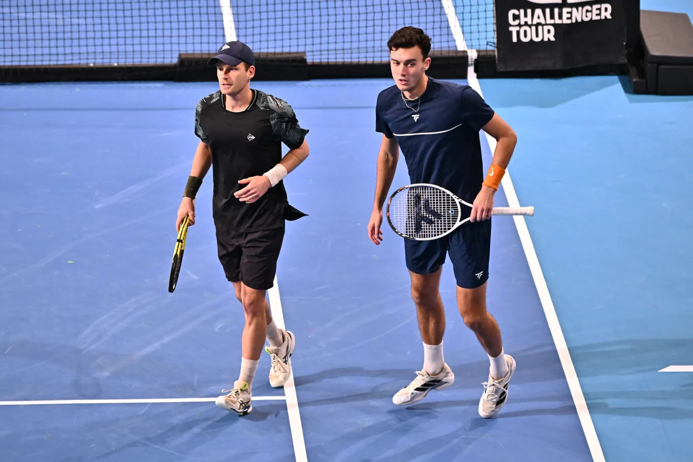 dwóch tenisistów w sportowych strojach znajduje się na niebieskim korcie podczas meczu deblowego, obaj trzymają rakiety tenisowe i są skoncentrowani na grze, w tle widoczna siatka oraz wyraźny napis 'Challenger Tour'