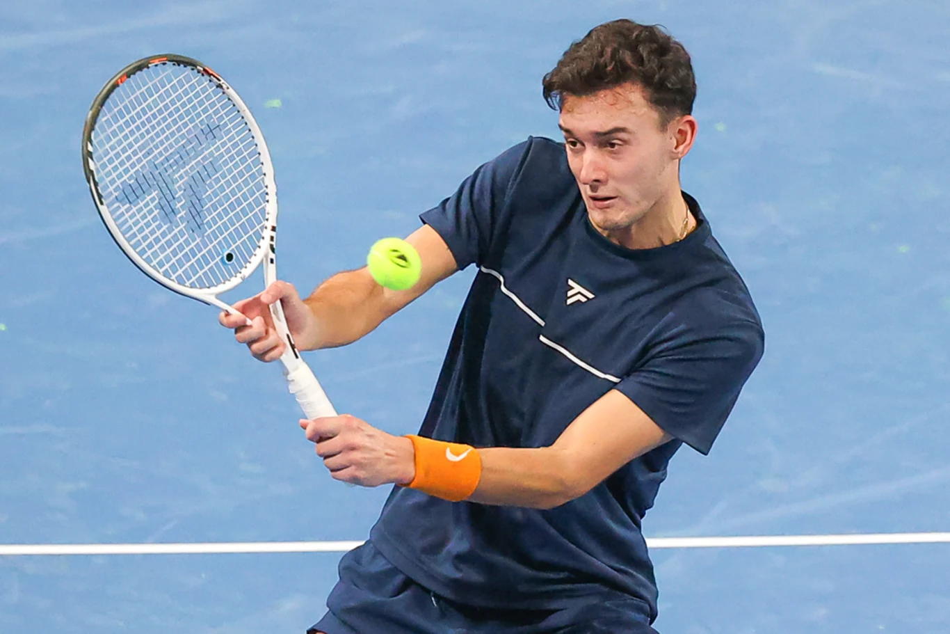 młody tenisista w granatowej koszulce odbija piłkę rakietą podczas dynamicznej wymiany na niebieskim korcie, skupiony na precyzyjnym ruchu i kontrolowaniu uderzenia