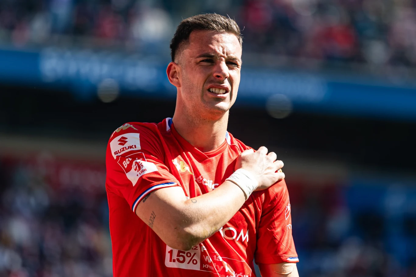 piłkarz w czerwonej koszulce trzyma się za lewe ramię z wyrazem bólu na twarzy, na tle stadionu pełnego kibiców