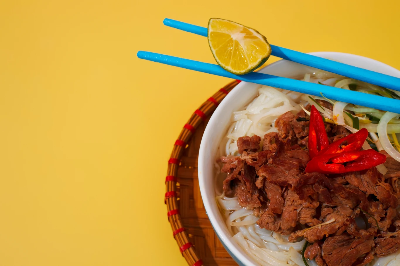 miska z makaronem ryżowym, kawałkami wołowiny, plasterkami czerwonej papryczki chili i dodatkiem świeżych warzyw, ułożona na plecionym podkładzie, z niebieskimi pałeczkami do jedzenia i kawałkiem limonki na żółtym tle