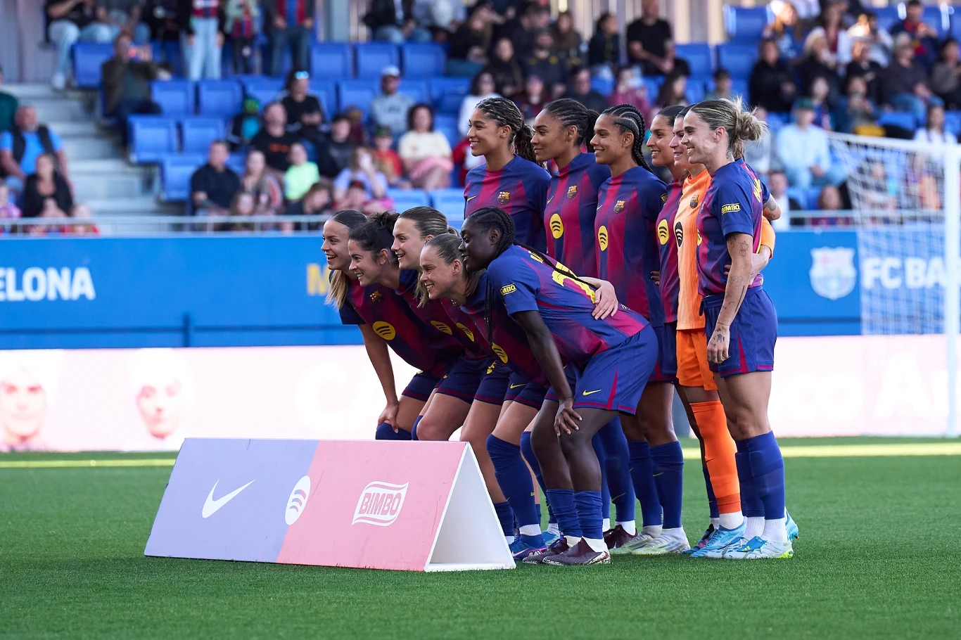 piłkarki drużyny FC Barcelona ubrane w klubowe stroje ustawione w dwóch rzędach do wspólnego zdjęcia na murawie stadionu, w tle widoczni kibice na trybunach oraz fragment bramki