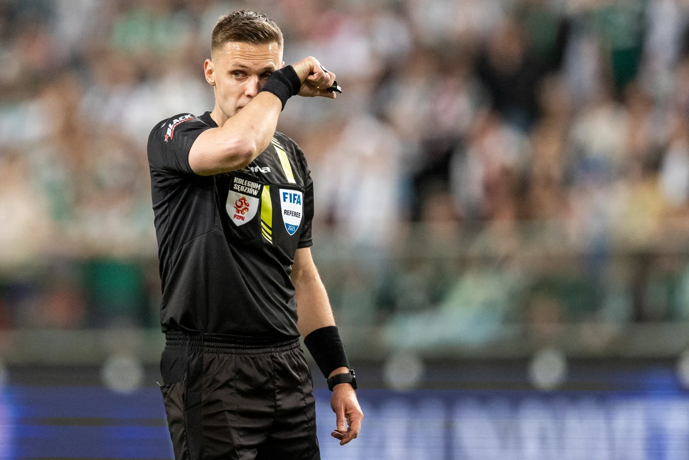 sędzia piłkarski w czarnej koszulce z odznaką FIFA oraz licznymi logotypami, wykonuje gest ręką przy twarzy na tle rozmytego stadionu wypełnionego kibicami