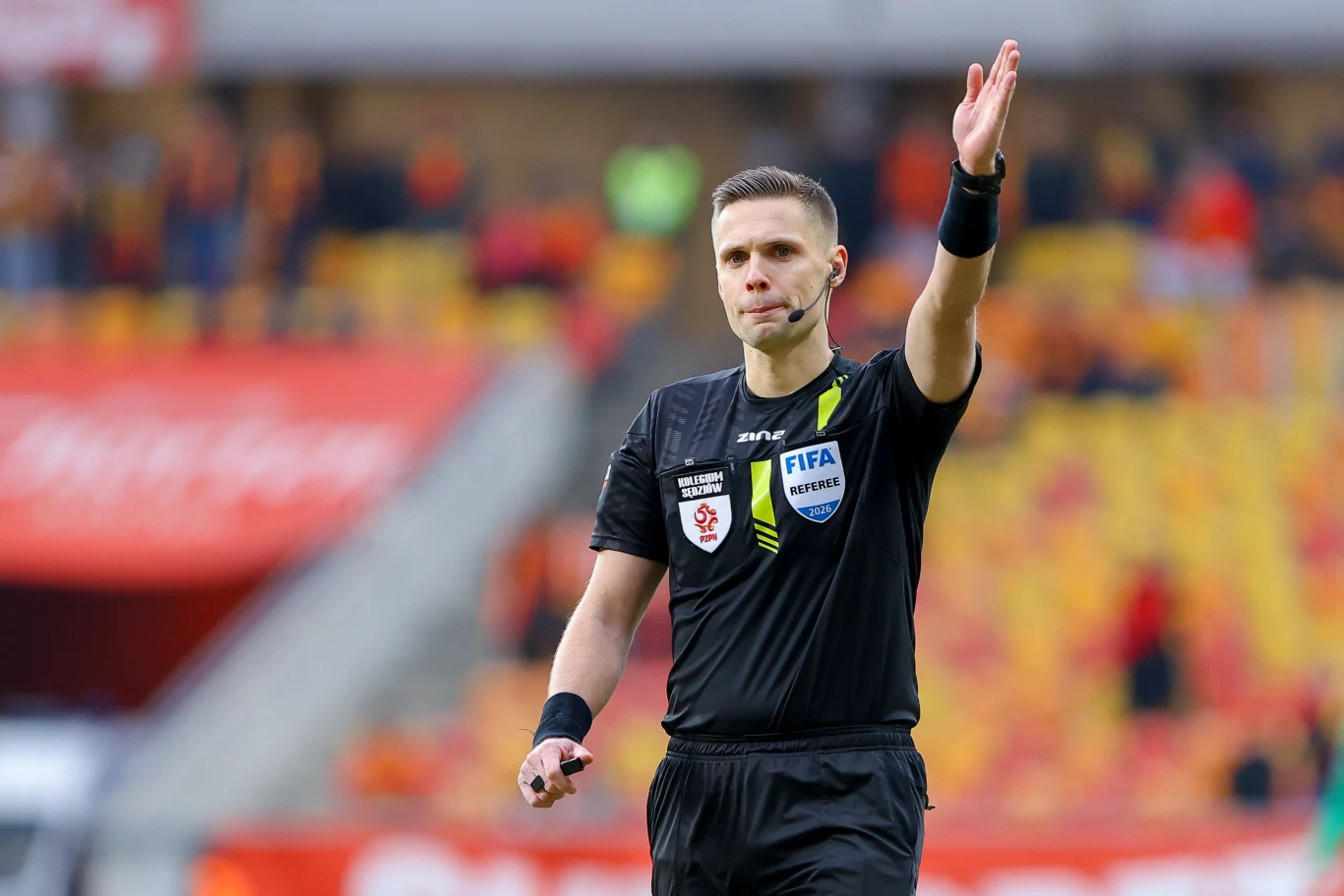 sędzia piłkarski w czarnym stroju z odznaką FIFA sygnalizujący decyzję podczas meczu, z uniesioną prawą ręką, na tle trybun wypełnionych kibicami