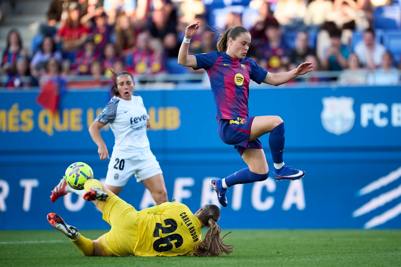 piłkarka w stroju Barcelony wykonuje skok nad bramkarką ubranej na żółto, która leży na murawie, w tle widać zawodniczkę drużyny przeciwnej i trybuny z kibicami