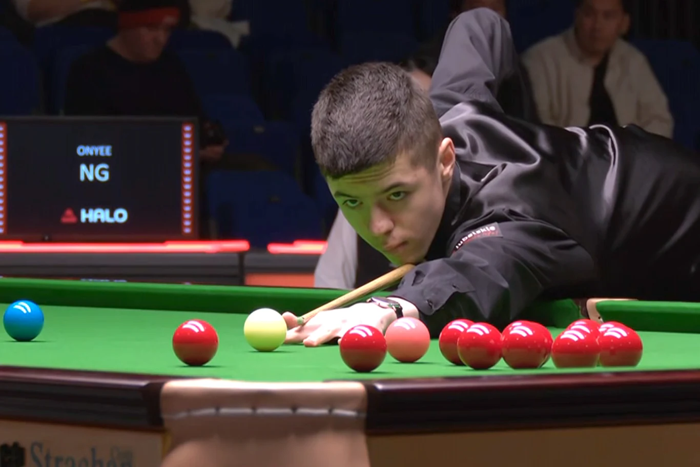 zawodnik w czarnym garniturze pochylony nad stołem bilardowym przygotowuje się do precyzyjnego uderzenia kijem bilardowym, koncentrując wzrok na białej bili pośród rozstawionych bili czerwonych i jednej żółtej