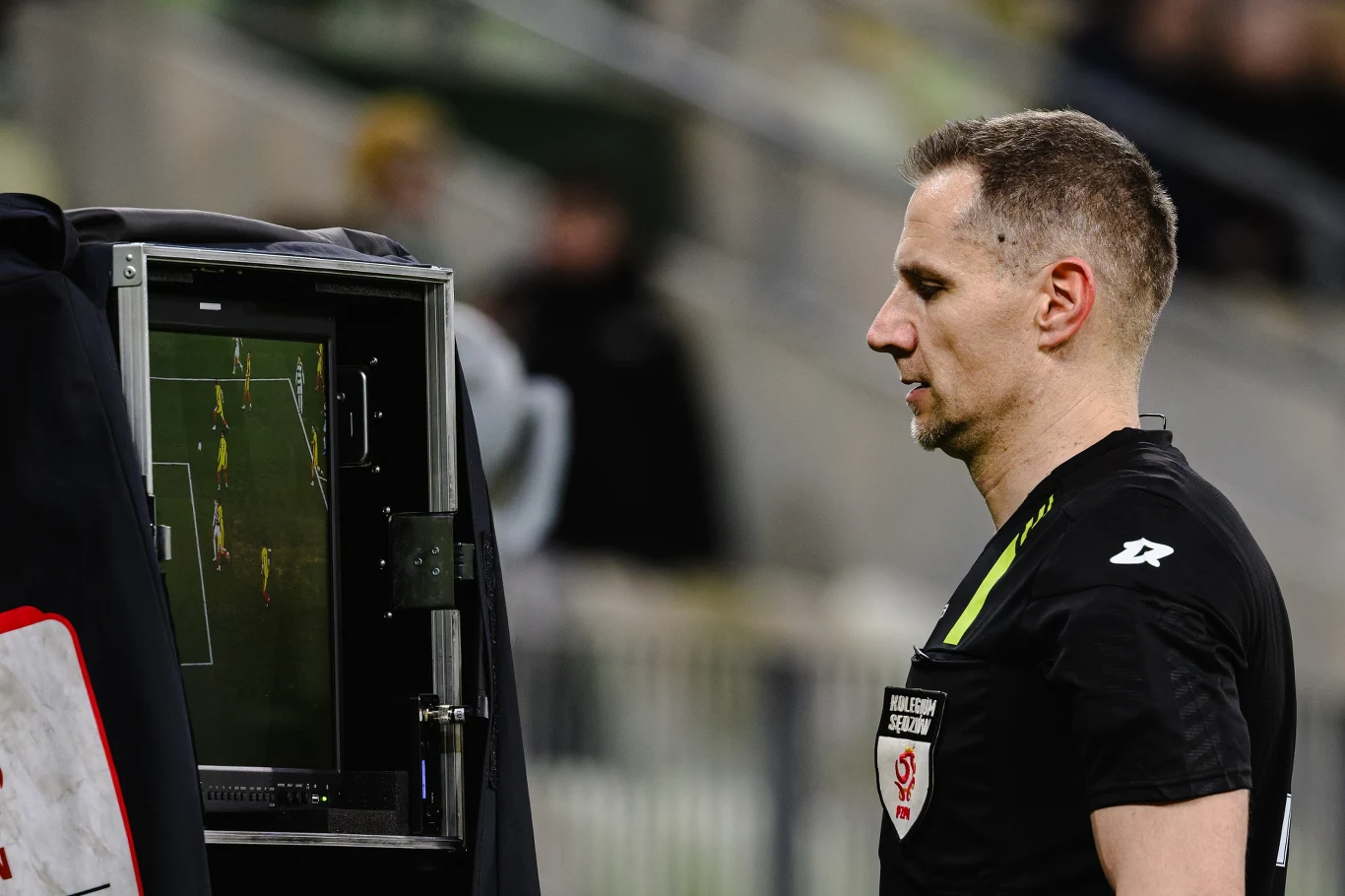 sędzia piłkarski analizuje powtórkę wideo na monitorze VAR podczas meczu, skupiając wzrok na ekranie z widocznym fragmentem boiska i zawodnikami
