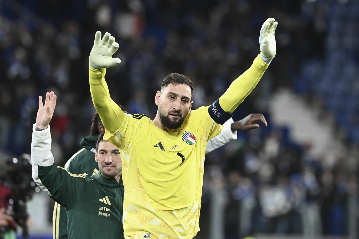 Bramkarz w żółtym stroju z uniesionymi rękami i opaską kapitana na ramieniu, witający lub żegnający się z kibicami na stadionie, w tle równie ubrani członkowie drużyny oraz rozmyte postaci na trybunach.