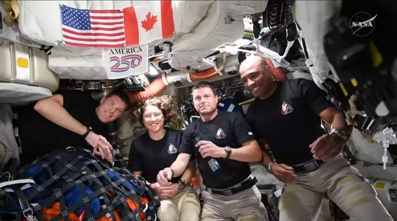 czterech astronautów w ciemnych koszulkach i jasnych spodniach znajduje się we wnętrzu stacji kosmicznej, w otoczeniu licznych urządzeń oraz worków zabezpieczonych siatką, na ścianie widoczne są flagi Stanów Zjednoczonych i Kanady oraz napis 'America 250'