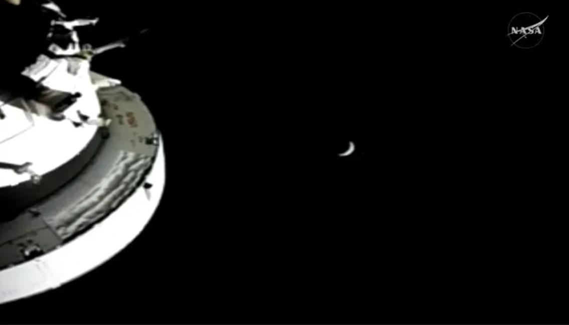 kapsuła kosmiczna zanurzona w czerni kosmosu z widocznym małym, półksiężycowym Księżycem w oddali; w prawym górnym rogu logo NASA