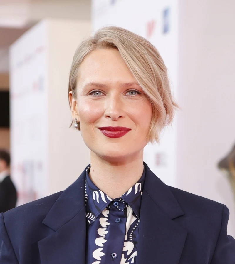 Agnieszka Żulewska Kobieta z krótkimi blond włosami i czerwoną szminką, ubrana w granatowy garnitur oraz koszulę z wzorem białych liści na ciemnym tle, stoi na tle jasnej ścianki z niewyraźnymi nadrukami i rozmytym sylwetkowym kształtem po prawej stronie.