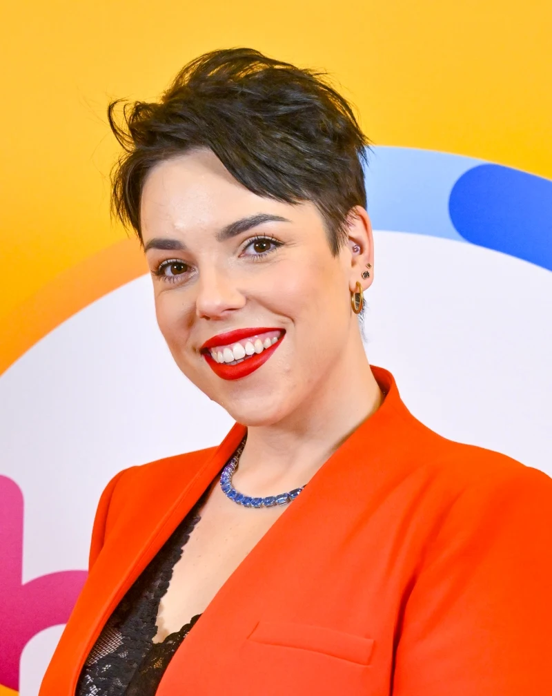 Maja Kumor Uśmiechnięta kobieta z krótkimi, ciemnymi włosami, ubrana w pomarańczową marynarkę oraz czarną, koronkową bluzkę, stojąca na tle w żywych kolorach z abstrakcyjnym wzorem w odcieniach żółci, bieli, niebieskiego i różu.