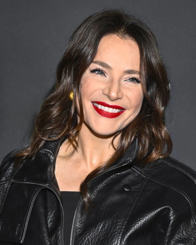 Anna Dereszowska kobieta z długimi, ciemnymi włosami, szeroko uśmiechnięta, ma intensywnie czerwone usta i nosi czarną, skórzaną kurtkę na tle jednolitej, ciemnej ściany