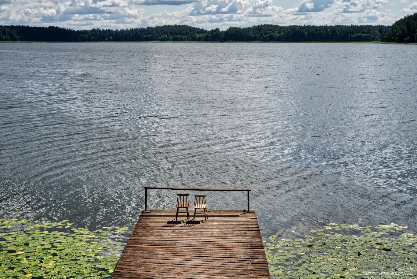 drewniany pomost z dwiema krzesłami skierowanymi w stronę spokojnego jeziora, otoczonego lasem, a przy brzegu unoszą się liście lilii wodnych