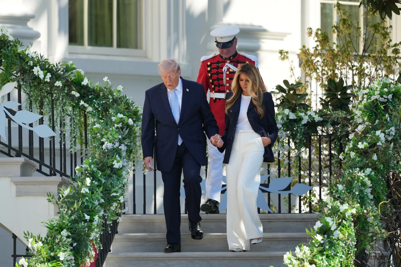Mężczyzna w granatowym garniturze i kobieta w białych spodniach oraz ciemnej marynarce schodzą razem po schodach udekorowanych zielonymi girlandami i białymi kwiatami, w tle stoi strażnik w paradnym mundurze z białą czapką.