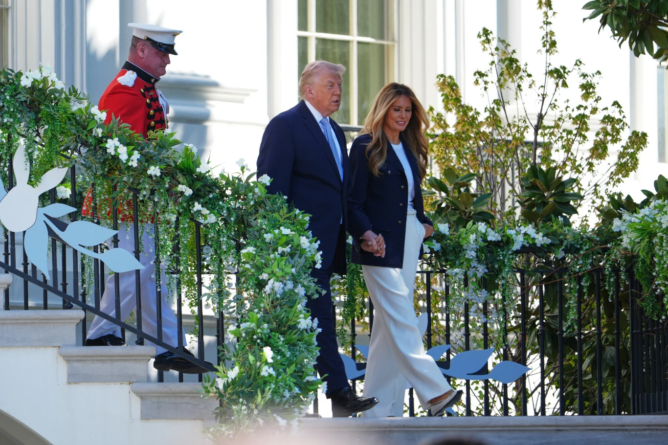 mężczyzna w garniturze i kobieta w jasnych spodniach oraz ciemnej marynarce trzymają się za ręce, idąc po schodach ozdobionych zielonymi i białymi dekoracjami kwiatowymi; z boku stoi żołnierz w czerwonym mundurze