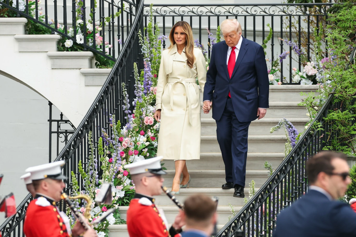 Para elegancko ubranych osób schodzi po zdobionych kwiatuszkami schodach na zewnątrz budynku, trzymając się za ręce, naprzeciwko nich grają członkowie orkiestry wojskowej w czerwonych mundurach.