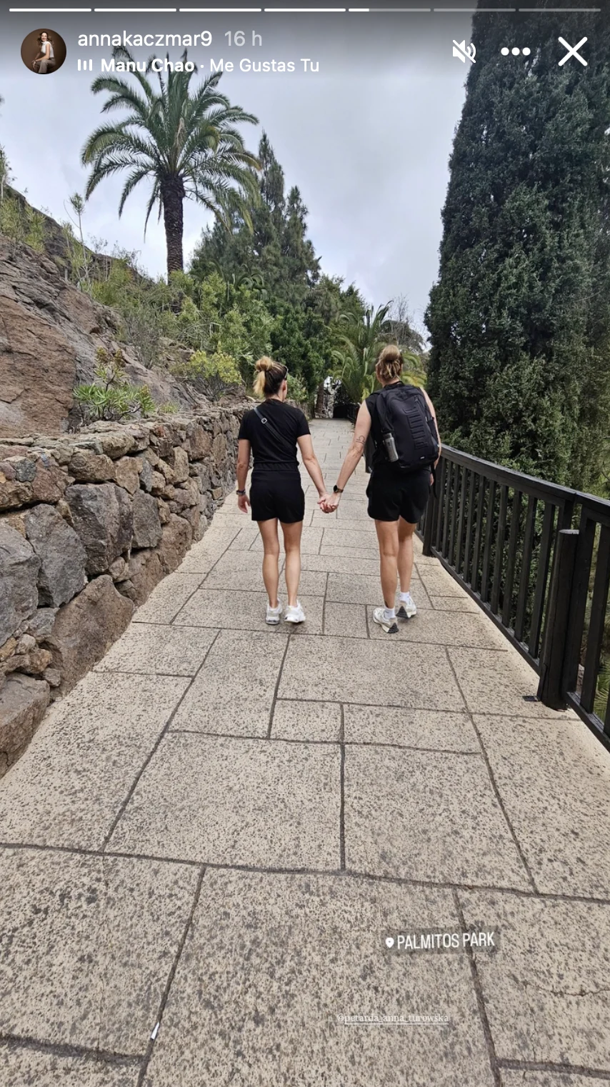 dwie osoby ubrane na czarno idą chodnikiem, trzymając się za ręce, otoczone zielenią z palmami i kamiennym murkiem po lewej stronie