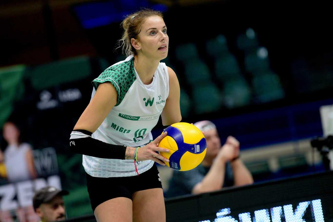 siatkarka w białej koszulce i czarnych spodenkach przygotowuje się do zagrywki, trzymając piłkę siatkową w obu rękach na hali sportowej