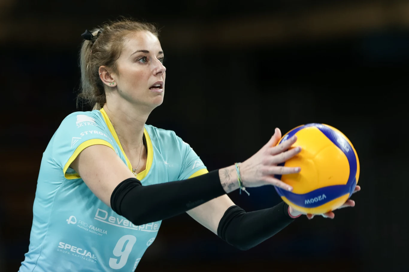 siatkarka w niebieskim stroju sportowym z numerem 9 przygotowuje się do zagrywki, skupiona trzymając piłkę do siatkówki obiema dłońmi