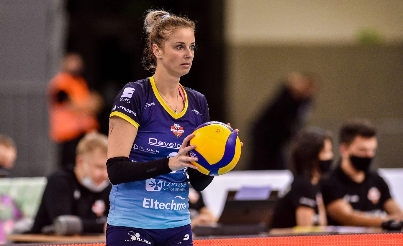 siatkarka w stroju sportowym przygotowuje się do serwisu, trzymając piłkę do siatkówki na tle hali sportowej, w tle widoczni sędziowie i osoby przy stoliku sędziowskim