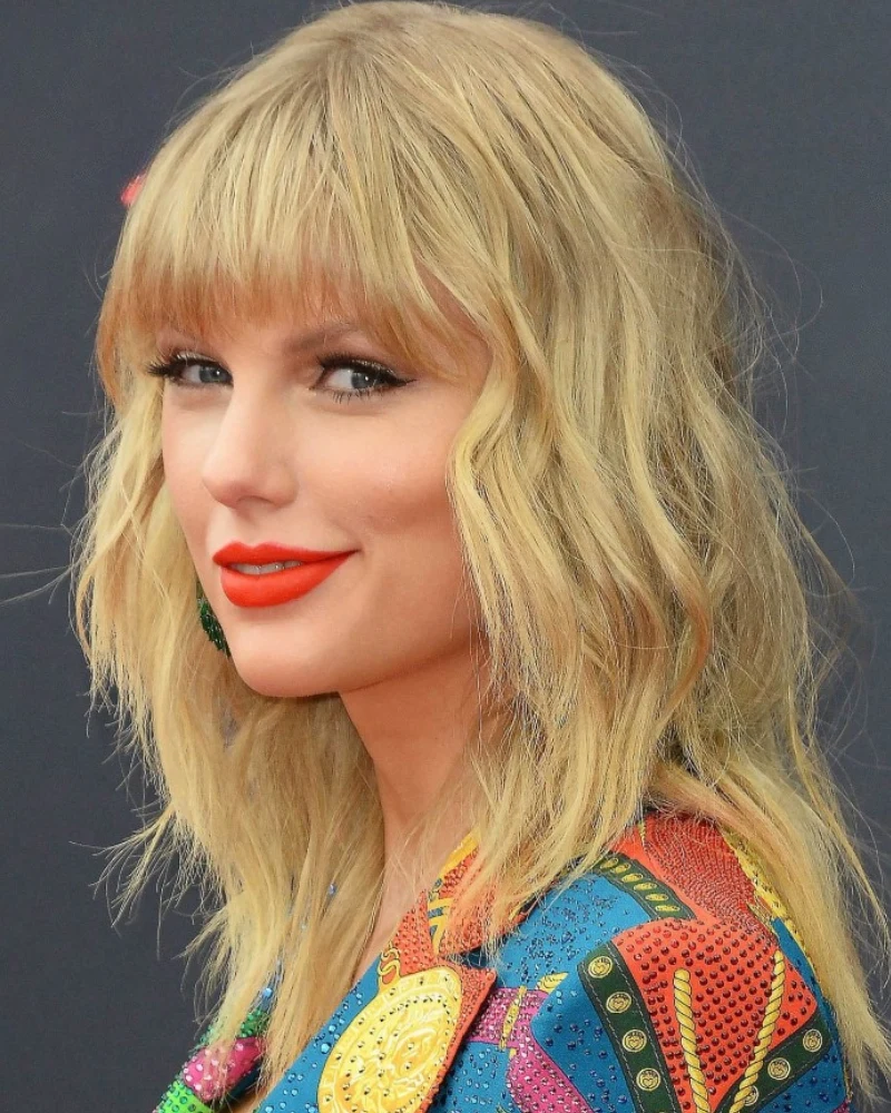 Taylor Swift Kobieta z jasnoblond falowanymi włosami, grzywką oraz wyraźnym makijażem, z ustami pomalowanymi na czerwono, ubrana w kolorową, wzorzystą marynarkę na tle ciemnego tła.