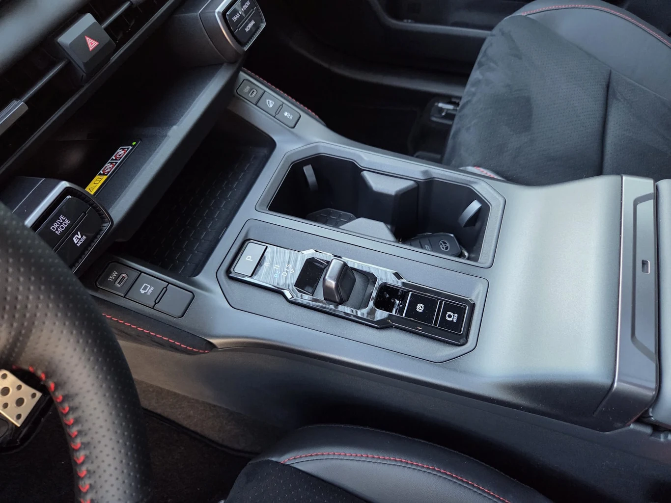 nowoczesna konsola środkowa samochodu z elektroniczną dźwignią zmiany biegów, dwoma uchwytami na napoje oraz przyciskami sterującymi systemami pojazdu, tapicerka z czarnego materiału ze sportowymi, czerwonymi przeszyciami