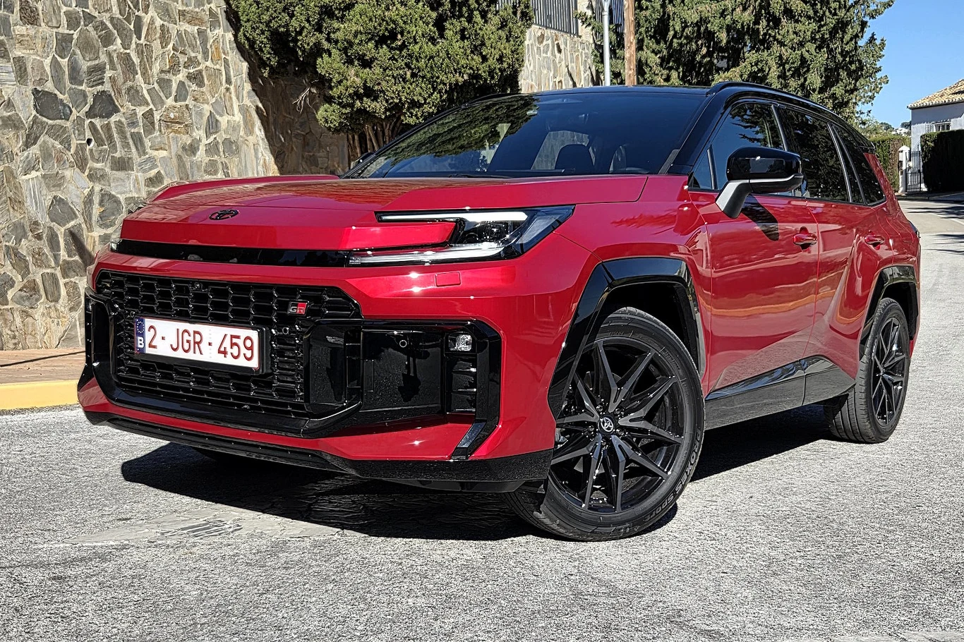 czerwony nowoczesny SUV z czarnym dachem i elementami karoserii, masywnym przednim grillem, agresywnym zderzakiem oraz czarnymi felgami, zaparkowany na ulicy w słoneczny dzień obok kamiennego muru i zielonych drzew