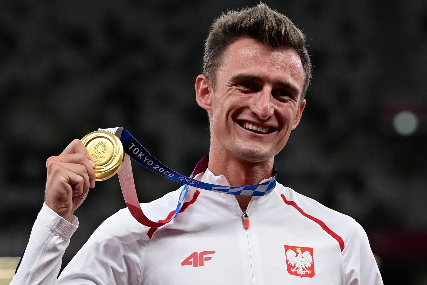 uśmiechnięty sportowiec w białym dresie z orłem na piersi trzyma w dłoni złoty medal olimpijski z napisem „Tokyo 2020”