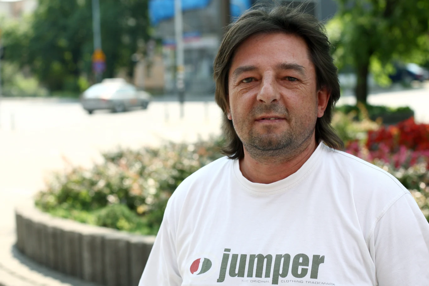 mężczyzna z ciemnymi włosami i zarostem w białej koszulce z napisem „jumper”, stojący na tle zieleni i kwiatów w miejskim otoczeniu za dnia