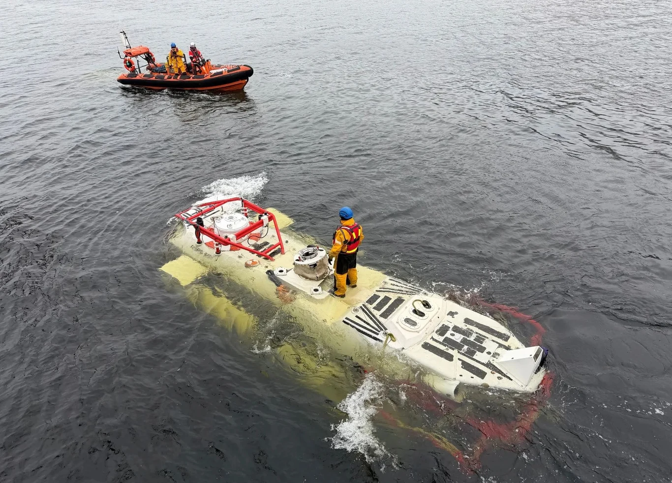 żółto-biały batyskaf unoszący się na powierzchni wody z osobą w kombinezonie i kasku na pokładzie, w tle niewielka pomarańczowa łódź ratownicza z kilkoma ludźmi na pokładzie