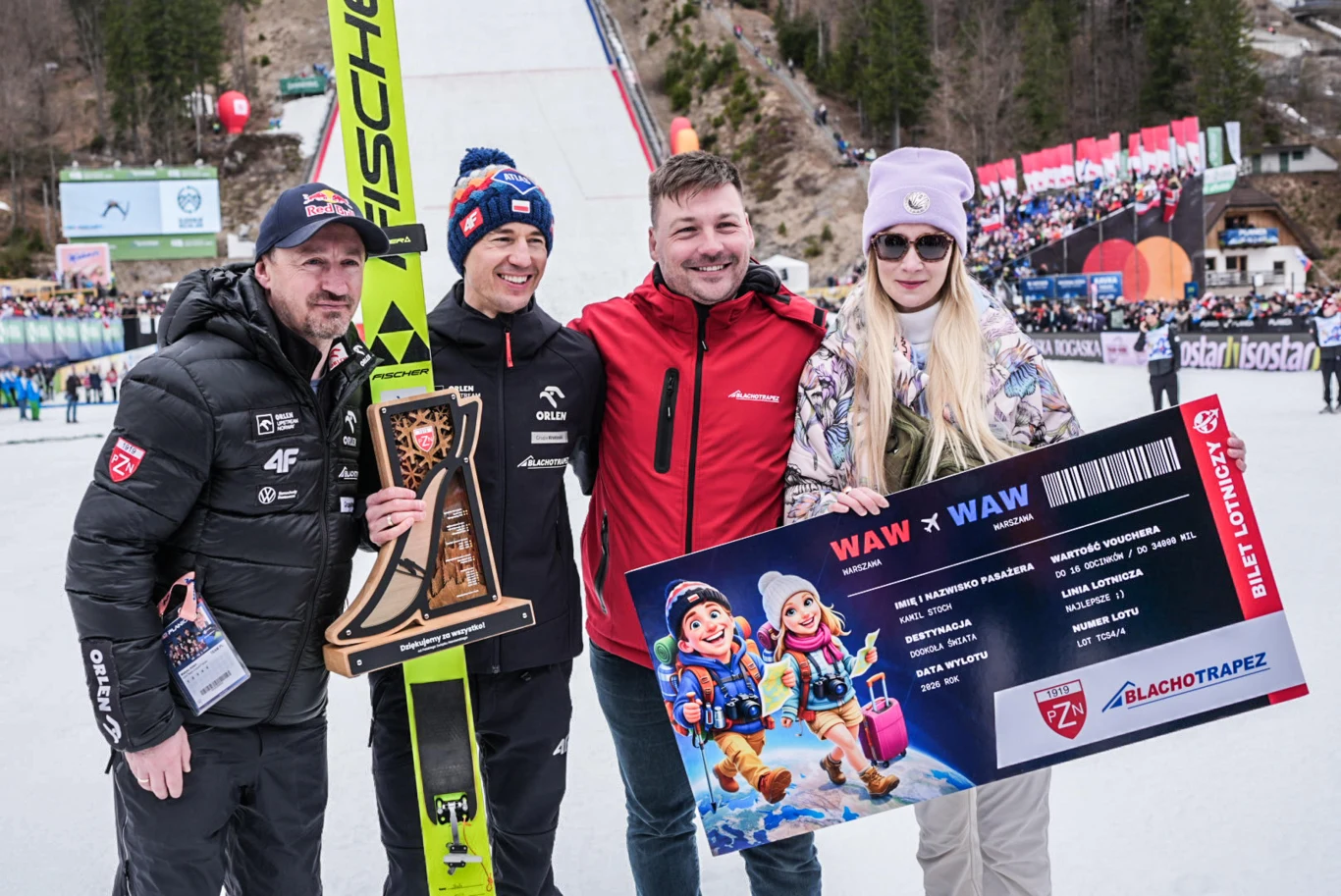 cztery osoby stojące razem na tle skoczni narciarskiej, jedna trzyma narty i trofeum, a inna trzyma dużą symboliczną tablicę przypominającą bilet lotniczy z grafiką maskotek, w tle widoczna publiczność oraz zimowa sceneria sportowa
