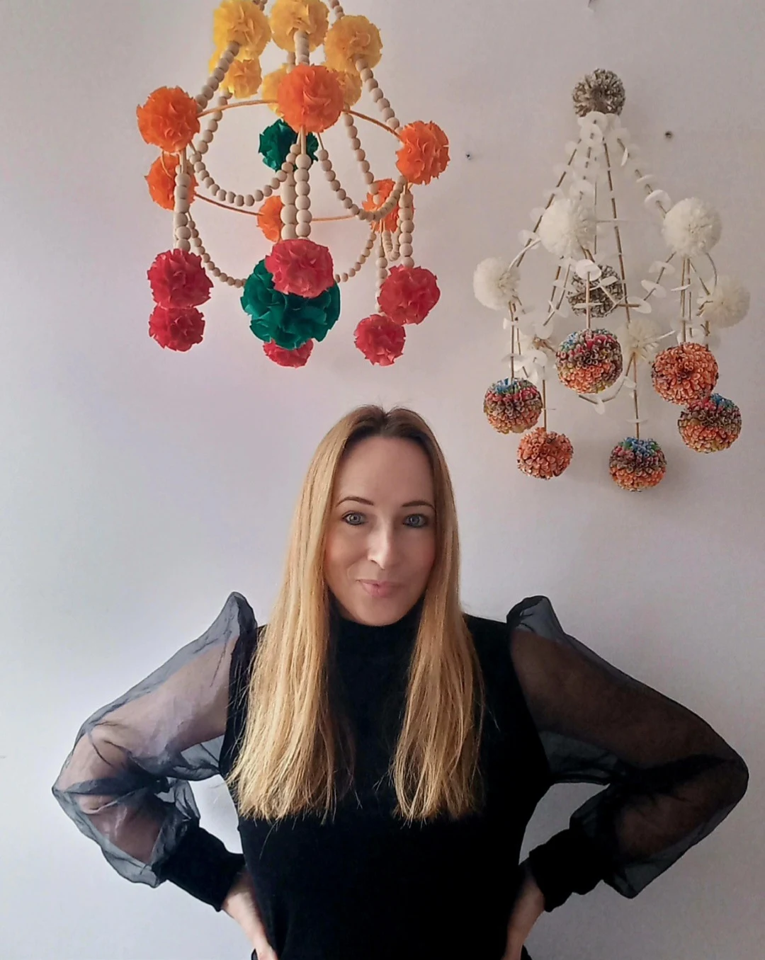 Kobieta z długimi blond włosami w czarnej bluzce z przezroczystymi rękawami stoi na tle dwóch kolorowych ozdobnych żyrandoli z pomponów zawieszonych na białej ścianie.