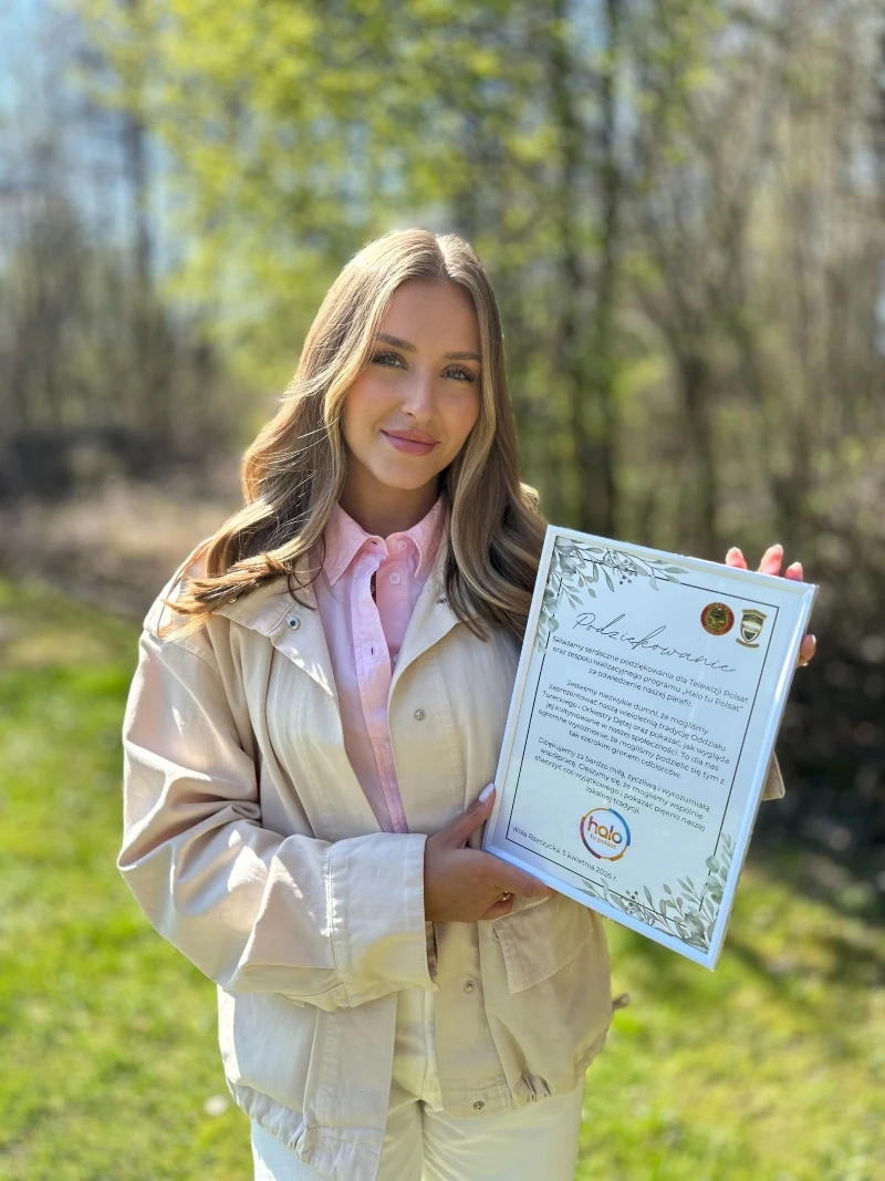 Klaudia Kroczek z podziękowaniami od mieszkańców młoda kobieta z długimi, jasnymi włosami ubrana w jasną kurtkę i różową koszulę, stojąca na tle zieleni w słoneczny dzień, trzymająca w rękach ozdobny dyplom z widocznym tekstem i logotypami