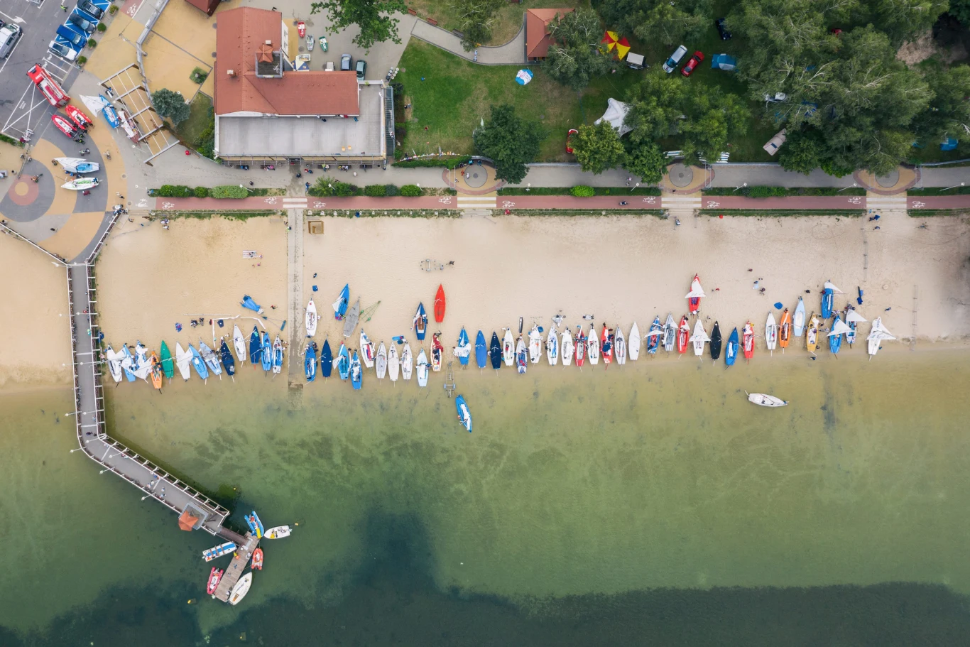 szereg kolorowych kajaków i łodzi ustawionych równolegle na piaszczystej plaży nad brzegiem jeziora, obok drewniany pomost wchodzący w wodę, w tle budynki i gęsta zieleń drzew