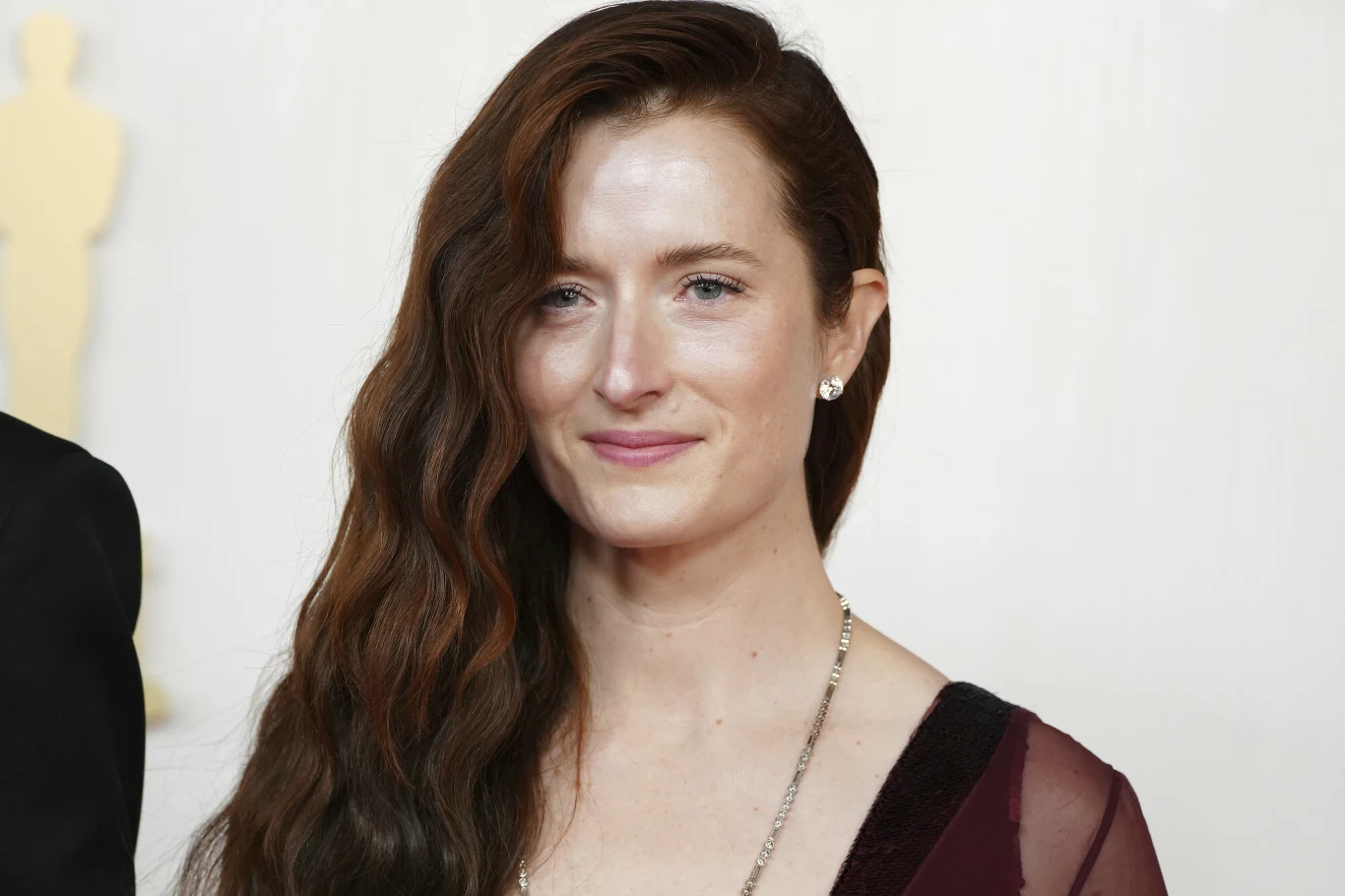 kobieta z długimi falującymi brązowymi włosami, ubrana w bordową suknię z dekoltem, subtelny makijaż i delikatna biżuteria w postaci kolczyków i naszyjnika, jasne tło podkreślające jej sylwetkę