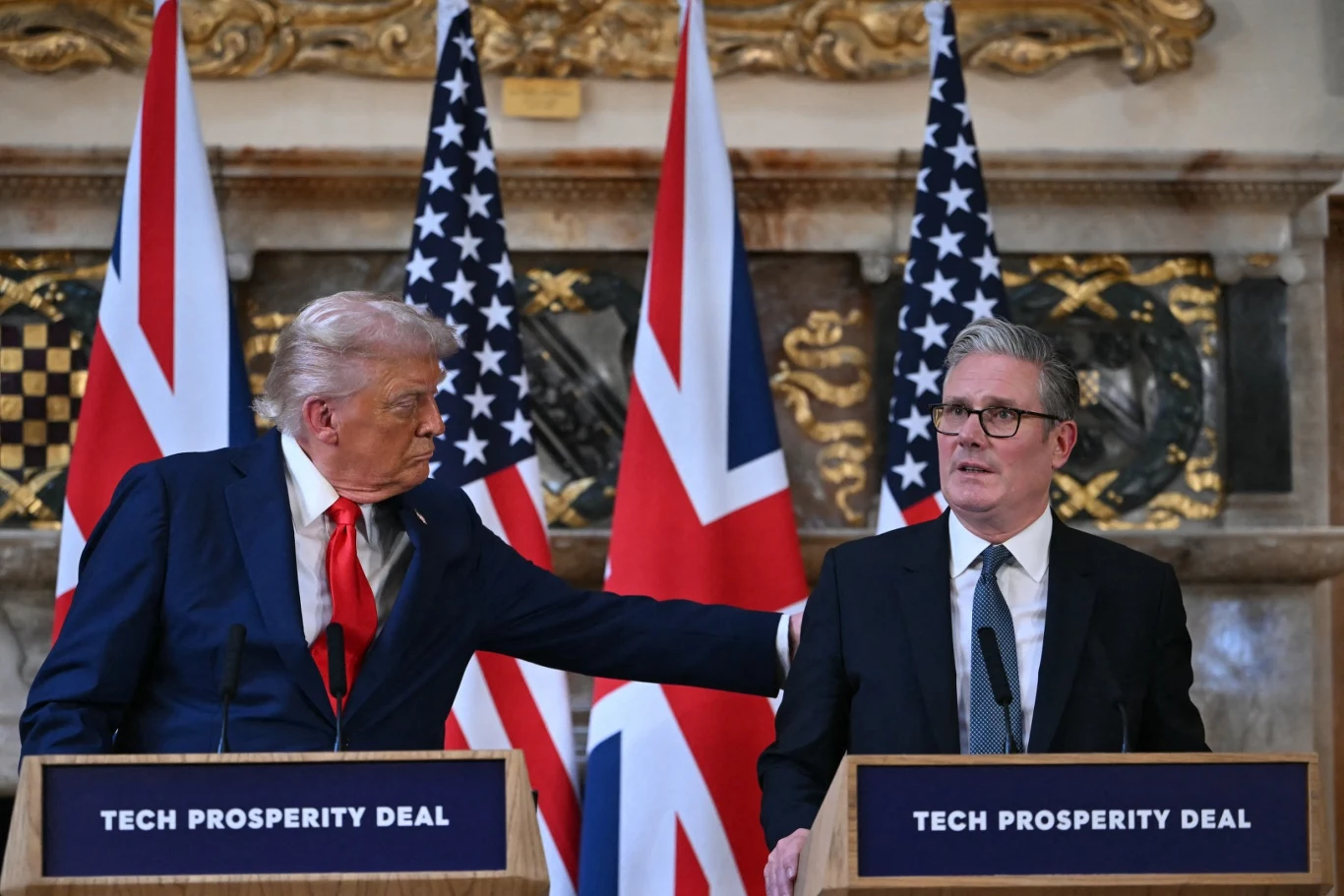 Dwóch mężczyzn stoi za mównicami z napisem "Tech Prosperity Deal" na tle flag Wielkiej Brytanii i Stanów Zjednoczonych; jeden z nich kieruje dłoń w stronę drugiego, obaj mają poważne wyrazy twarzy i są elegancko ubrani w garnitury.