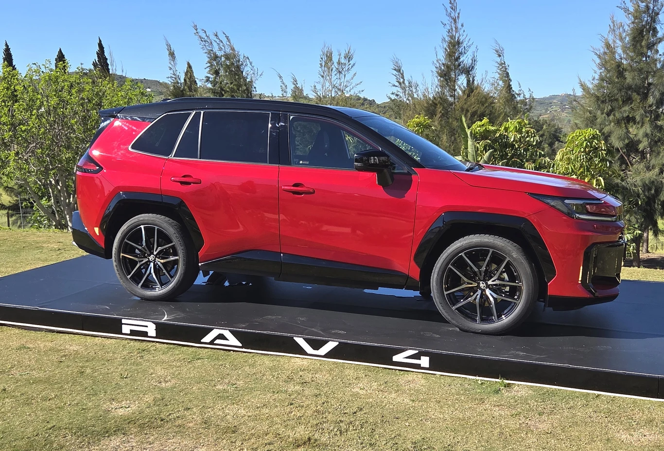 Czerwony SUV Toyota RAV4 z czarnym dachem i dużymi, sportowymi felgami stoi na podwyższonej, czarnej platformie na tle zielonych drzew i nieba.
