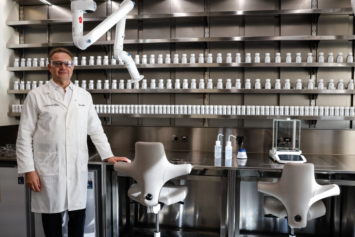 mężczyzna w białym fartuchu laboratoryjnym i okularach ochronnych stoi w nowoczesnym laboratorium z metalowymi blatami, dwoma białymi krzesłami, ramieniem robota laboratoryjnego oraz licznymi jednakowymi butelkami ustawionymi w równych rzędach na półka...