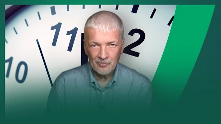 mężczyzna o siwych włosach, ubrany w koszulę, umieszczony na tle dużej tarczy zegara z widocznymi godzinami 11 i 2