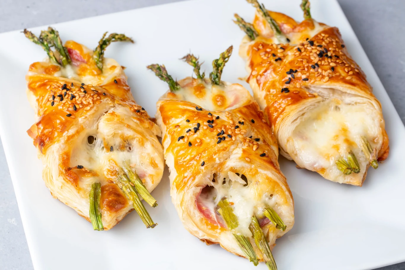 Baked green asparagus with ham and cheese in puff pastry sprinkled with sesame seeds and green basil leaves. Przekąski z ciasta francuskiego z zielonymi szparagami, serem i szynką, posypane sezamem, na białym talerzu.