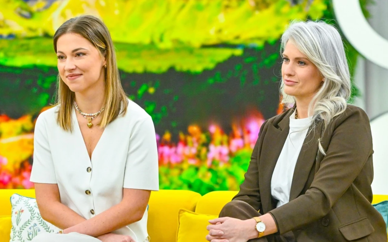 Dwie kobiety siedzące obok siebie na żółtej sofie, jedna w jasnej sukience, druga w ciemnym garniturze, w tle kolorowa, rozmyta dekoracja ścienna.