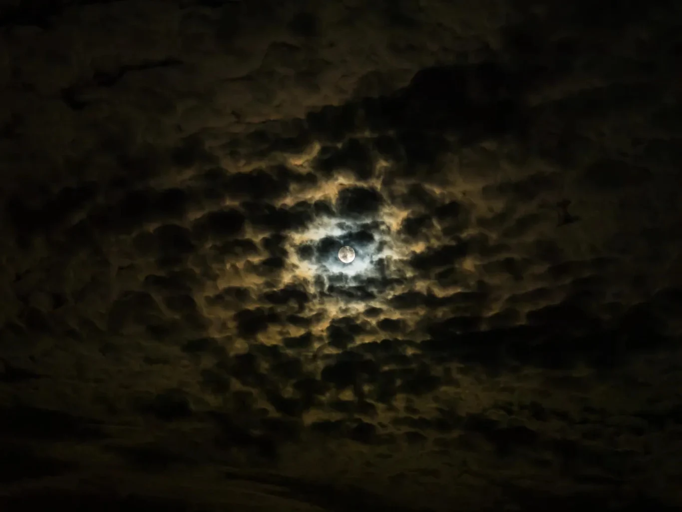 jasny księżyc otoczony gęstą warstwą ciemnych, rozproszonych chmur, przez które przebijają się promienie światła tworząc tajemniczy, dramatyczny efekt