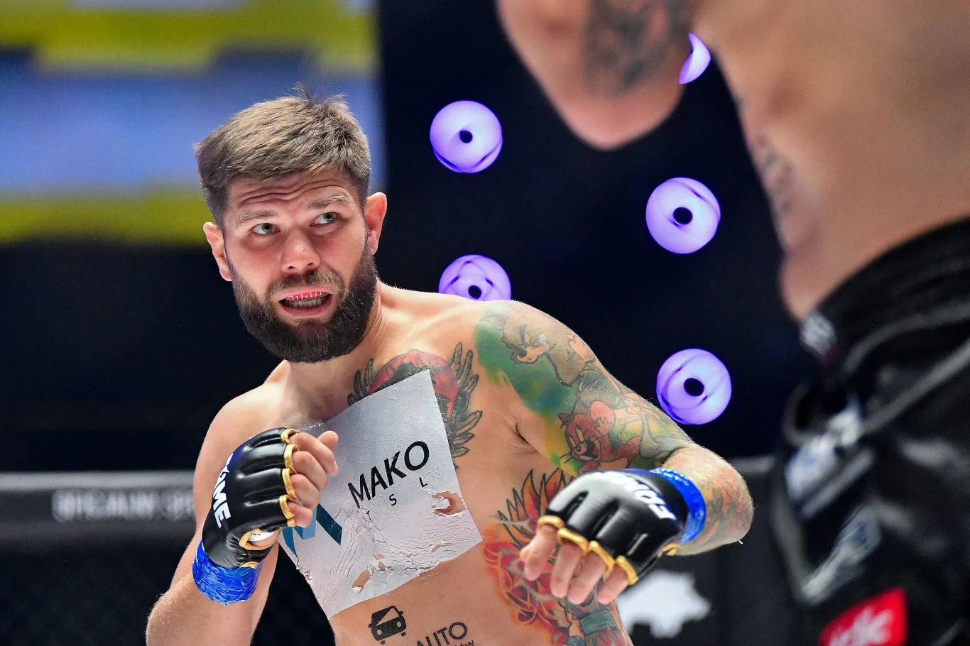 Zawodnik MMA z tatuażami na ramieniu i torsie przygotowujący się do ataku podczas walki, skupiony wzrok skierowany na rywala, obaj zawodnicy w rękawicach, dynamiczna atmosfera pojedynku na arenie sportowej.