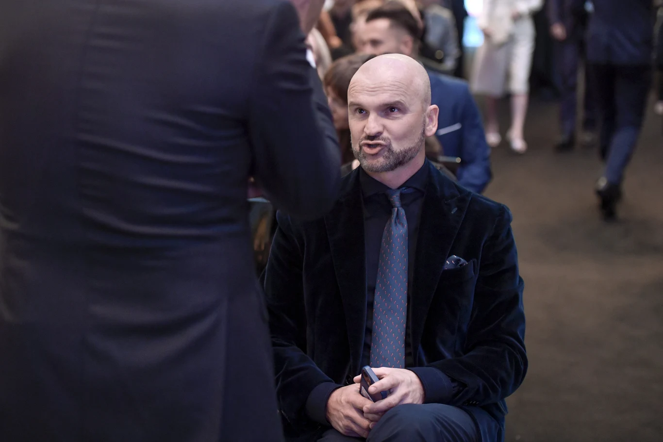 Łysy mężczyzna z brodą, ubrany w elegancki, granatowy garnitur i krawat, siedzi na krześle podczas rozmowy z inną osobą, która stoi przed nim tyłem do obiektywu, w tle widać inne osoby ubrane formalnie.